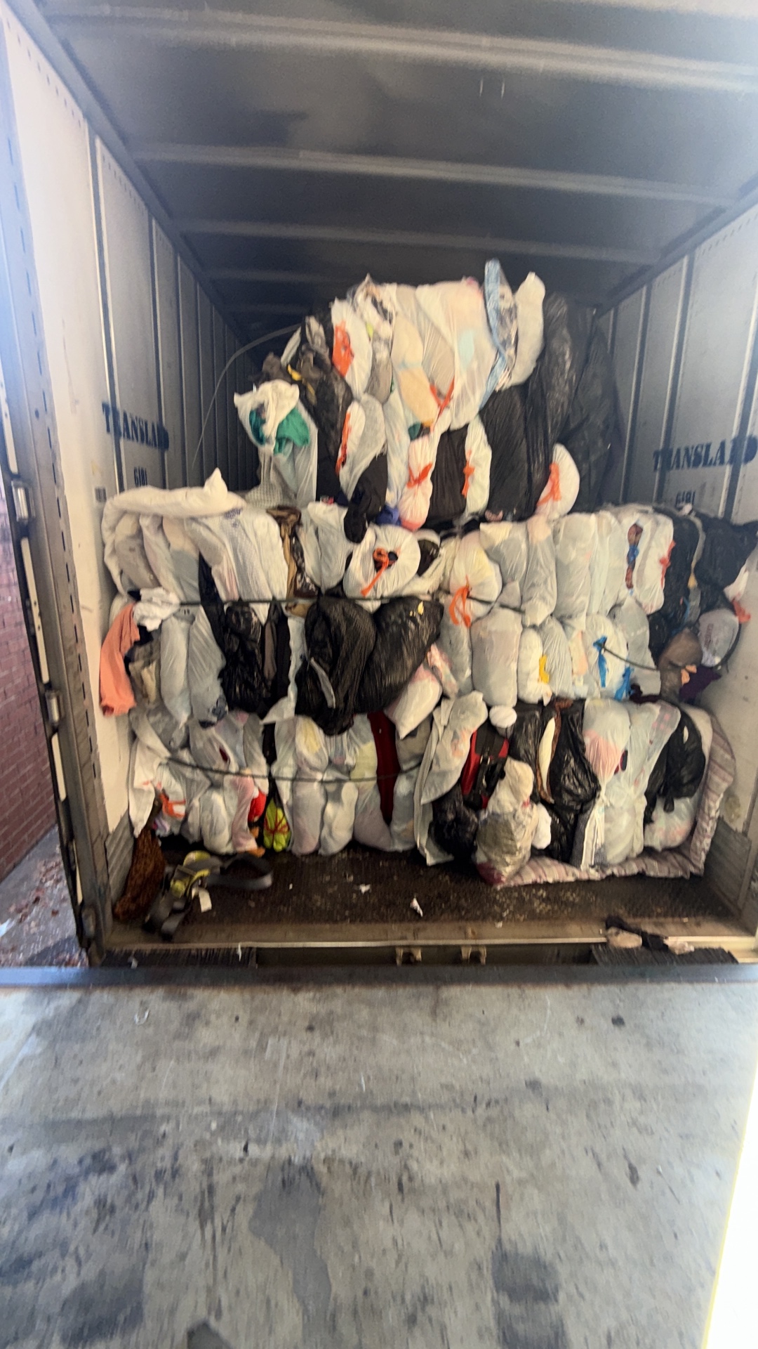 Workers loading bulk used clothing in front of stacks of compressed premium credential clothing bales inside a recycling warehouse